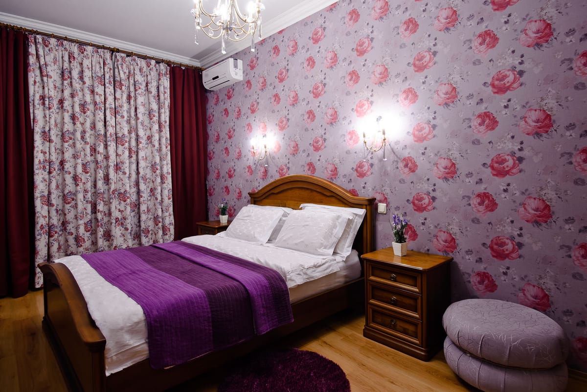 Bedroom with rose-patterned wallpaper, a purple runner across the bed and a floor cushion