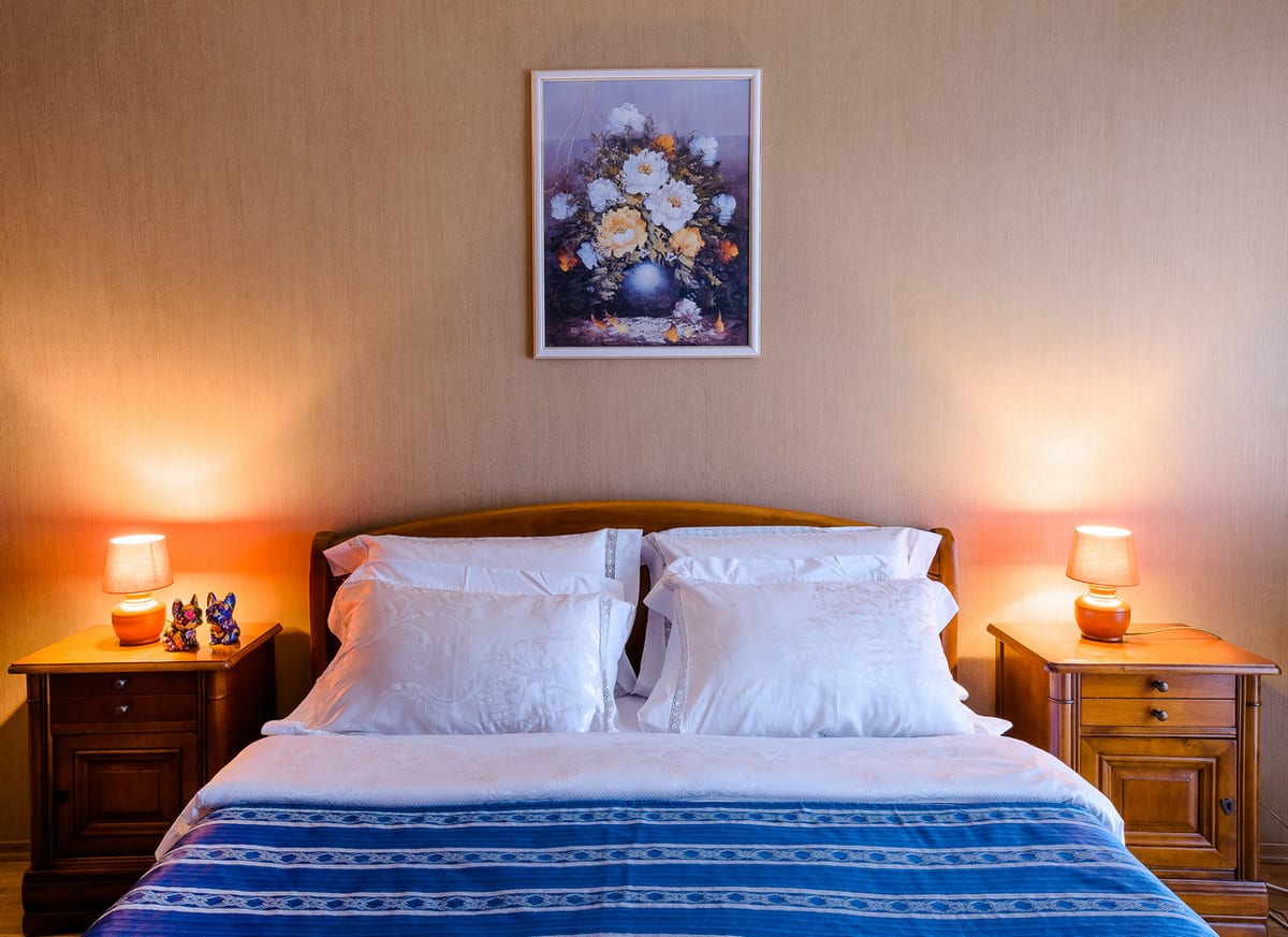 Double bed between two lit bedside lamps beneath a framed floral still life