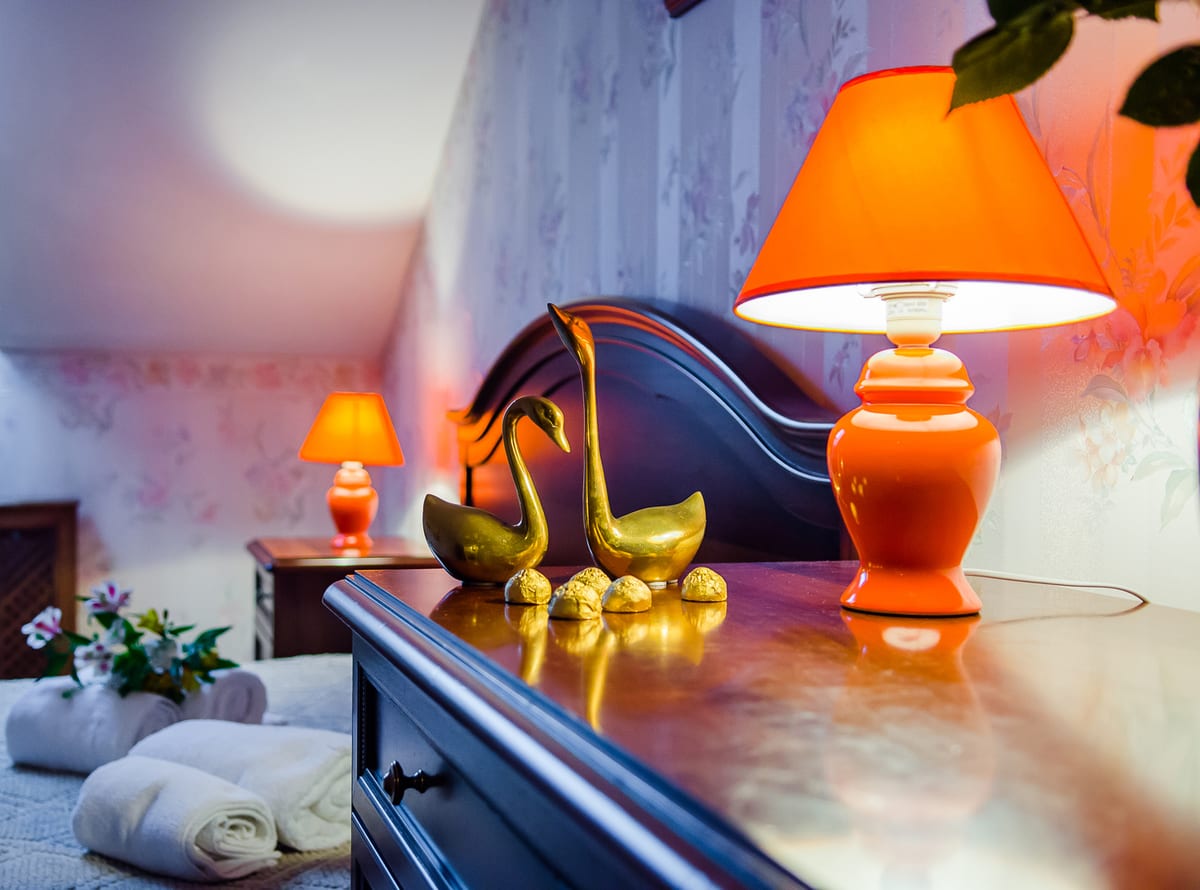 Bedside table with an orange lamp, brass swan ornaments and rolled towels on the bed