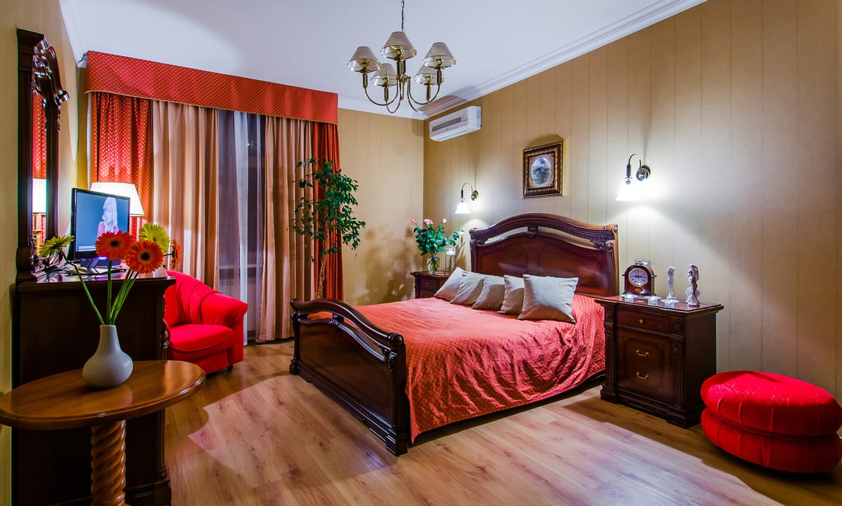 Bedroom with a red bedspread, a carved bed, a chandelier and a red velvet pouffe