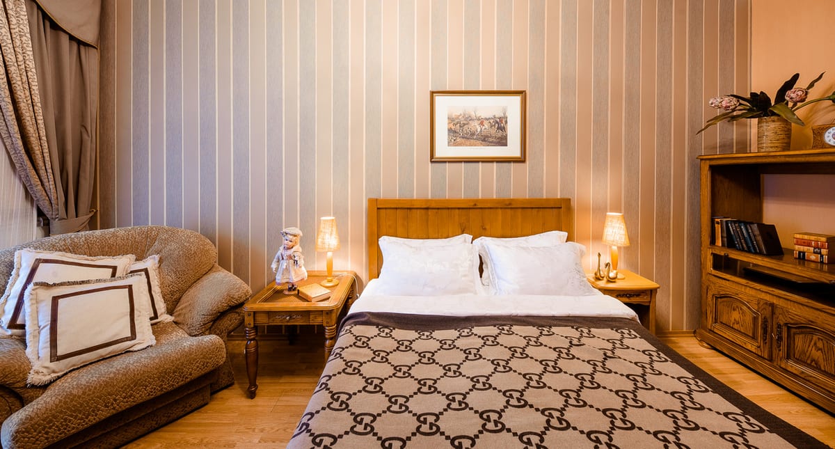Bedroom with striped wallpaper, an armchair piled with cushions and a bookcase