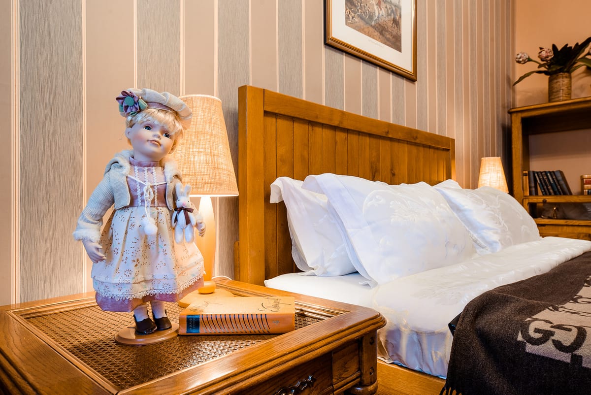 A porcelain doll and a lamp on the table beside a made-up bed