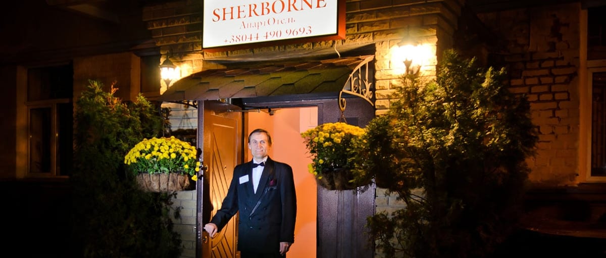 Hotel entrance at night, the lit SHERBORNE sign and a doorman in the open doorway