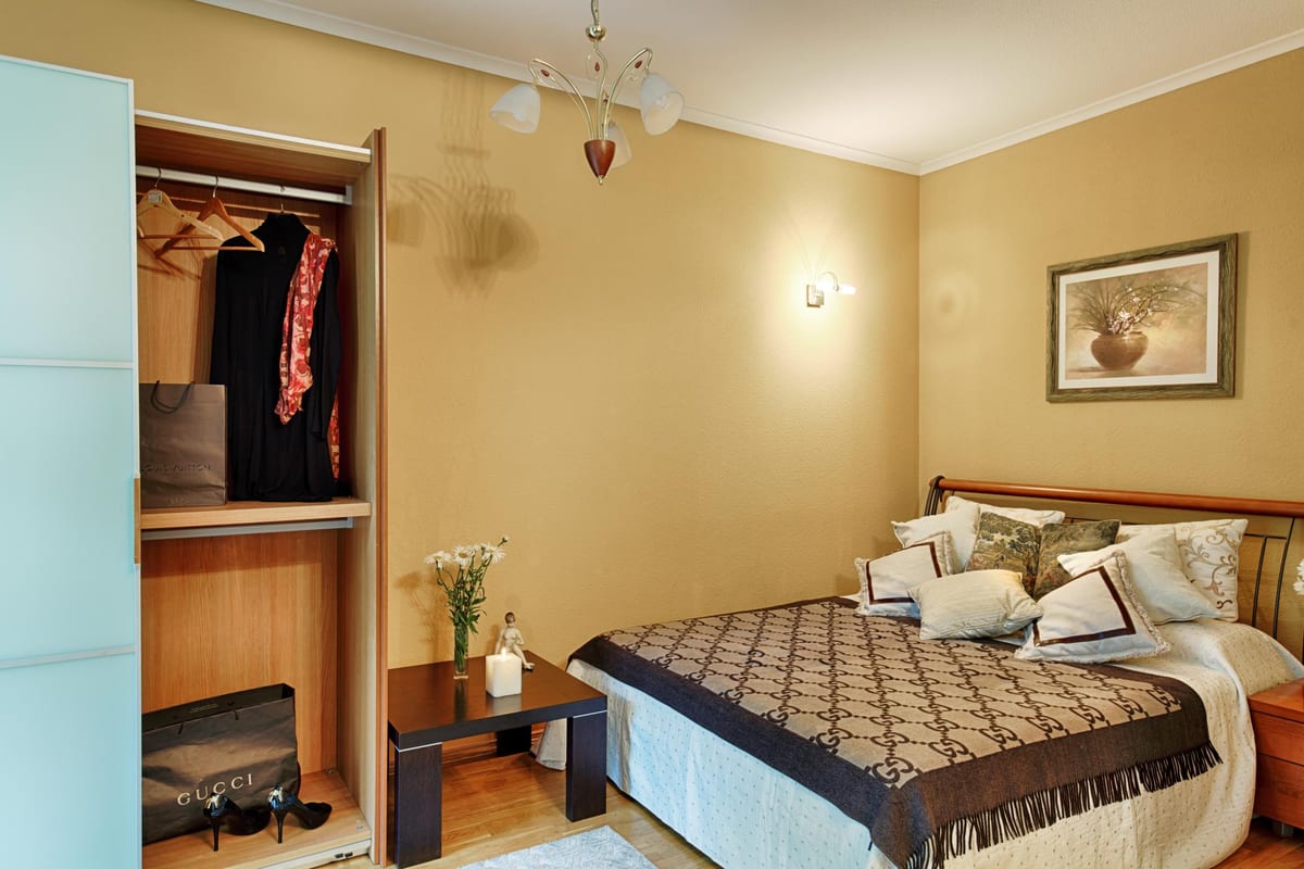 Bedroom with an open wardrobe, ochre walls and a monogrammed throw over the bed