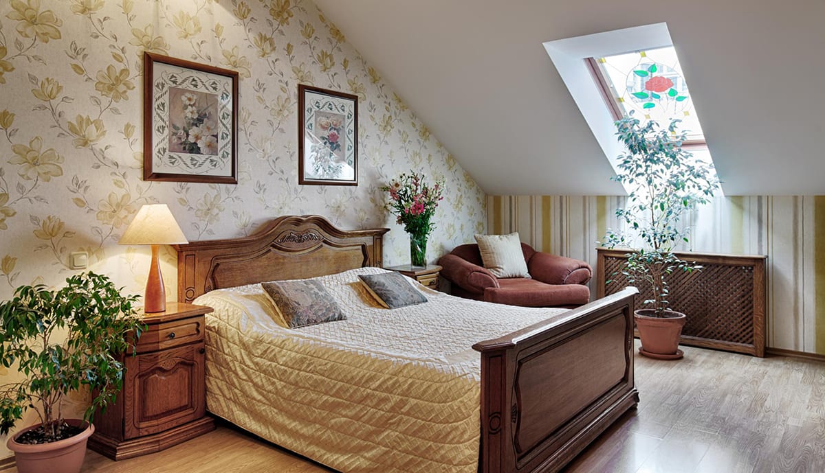 Floral mansard bedroom seen wide, armchair and potted tree under the sloping ceiling